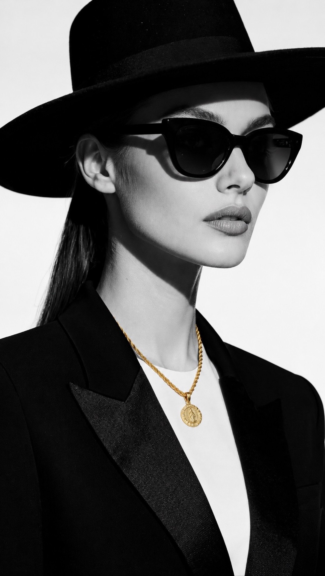 Modelo en blanco y negro con sombrero y gafas oscuras luciendo completa la cadena cordón franciscano con el dije de San Benito en oro 18K, la única pieza en color
